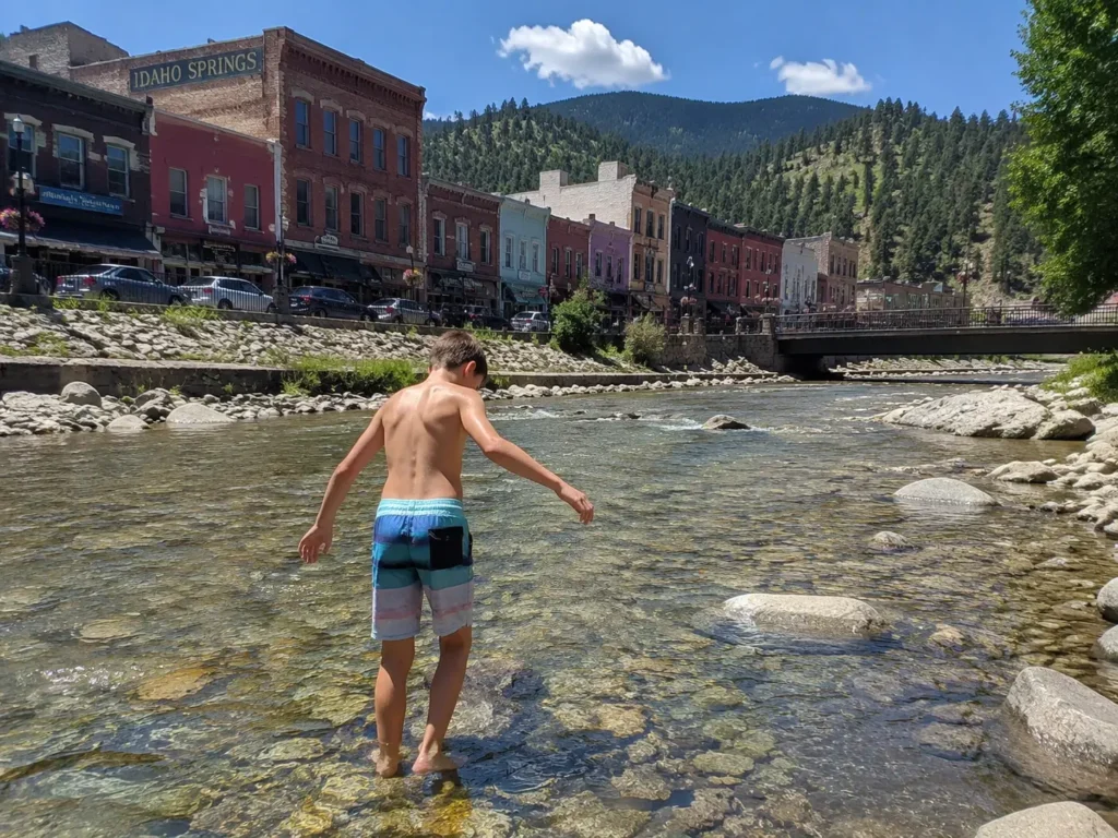 Boy wading in Clear Creek with Idaho Springs historic downtown buildings visible on the bank